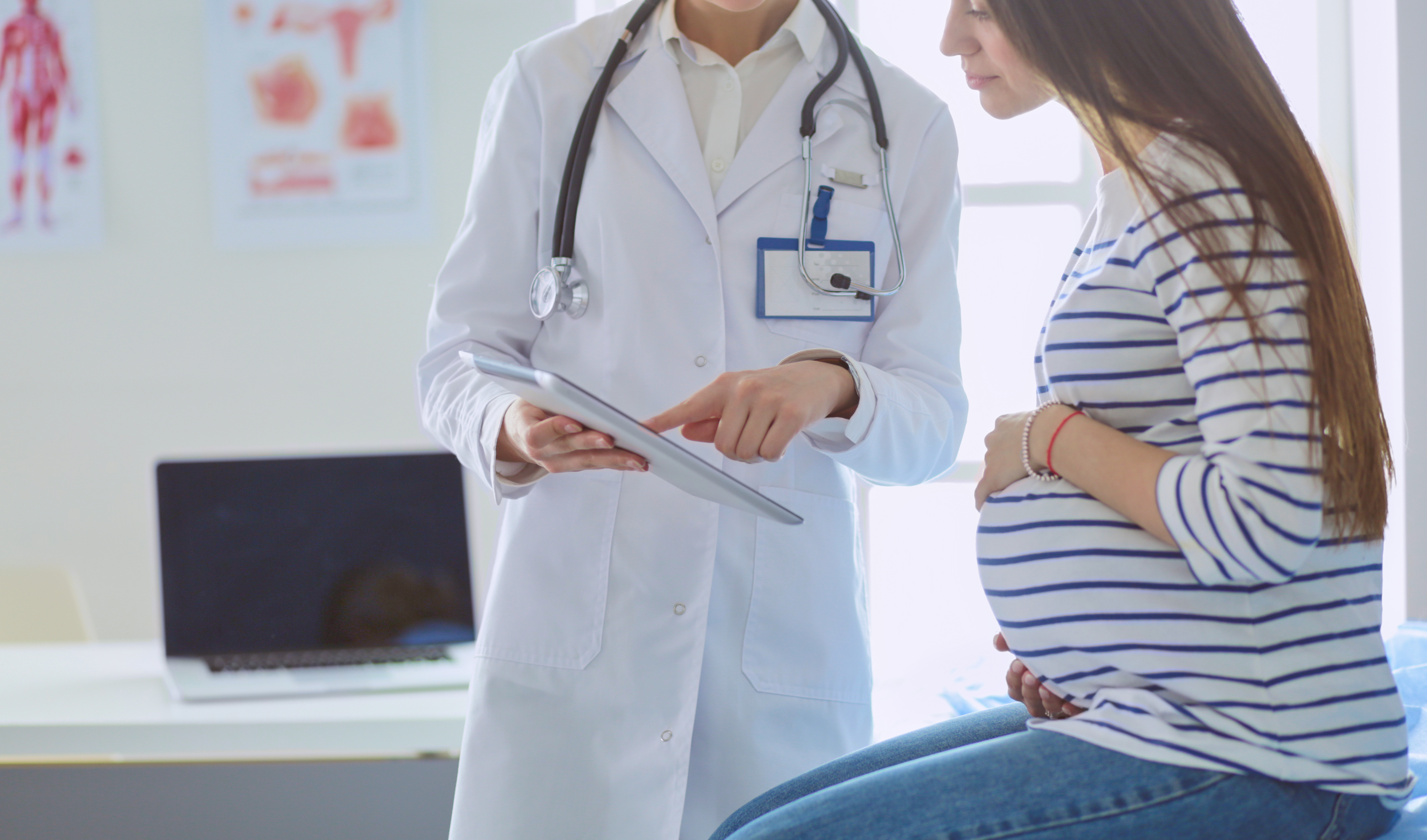 Pregnant woman sitting on exam table talking with doctor holding tablet