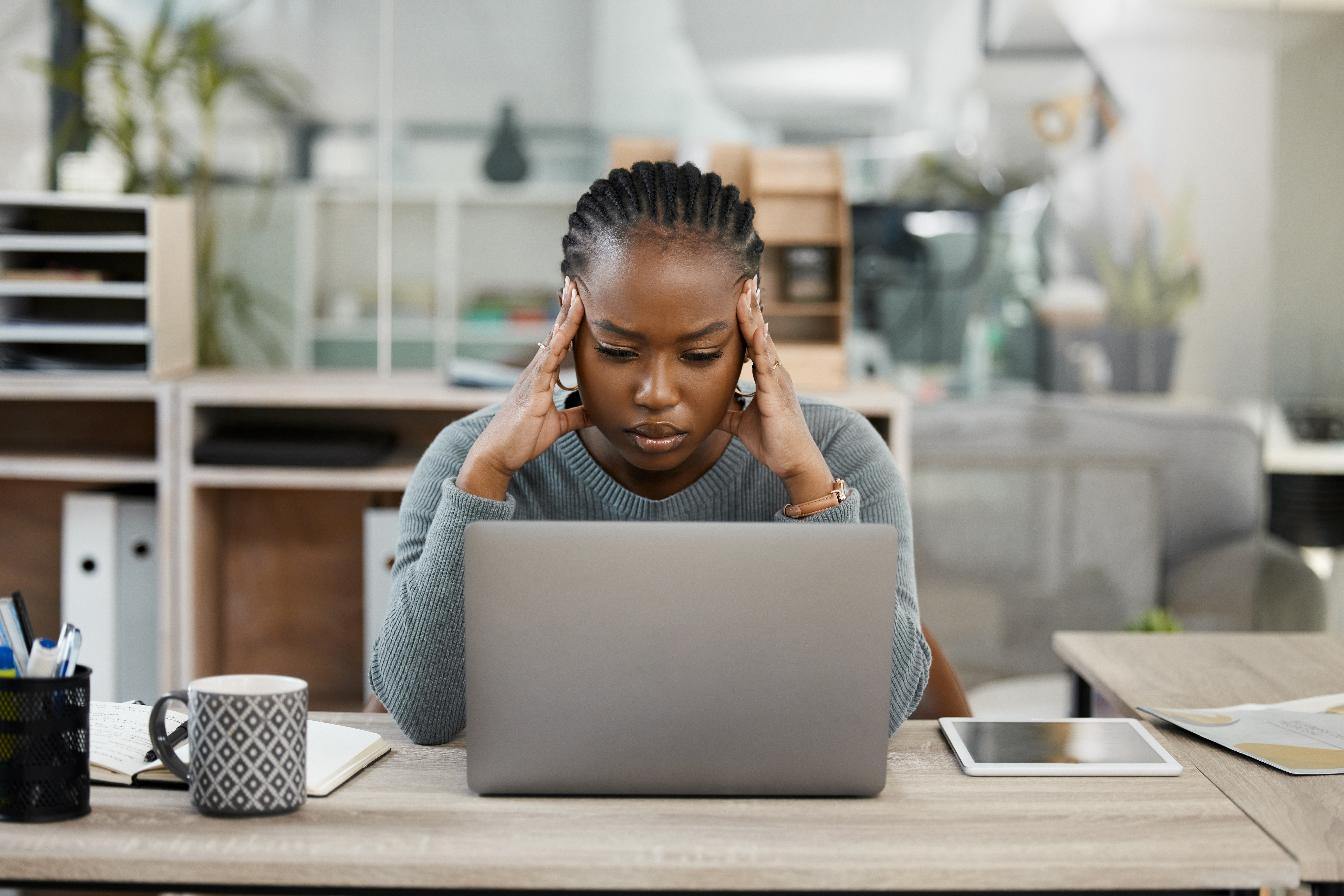 A woman looks down at her laptop in frustration. It's daytime in what looks like a home office.