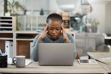 A woman looks down at her laptop in frustration. It's daytime in what looks like a home office.