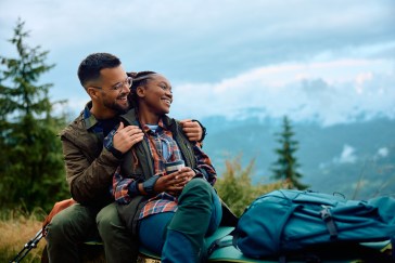 A Black couple sits on a bench in hiking clothes at the top of a mountain, smiling at the view.
