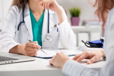 A female doctor uses a pen to point to something on a chart to a patient sitting across from her.