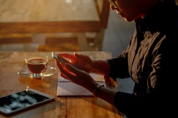 A woman sitting outside during sunset stares down at her phone; a cup of tea and an iPad are on the table.