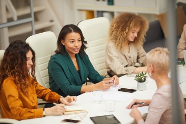 A group of women at a conference table with tablets, phones and papers.