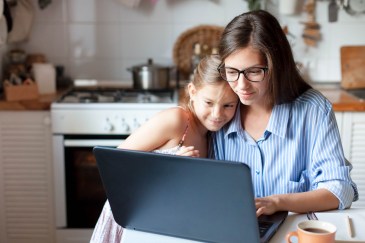 A mom hugs her young daughter while looking at her laptop at the kitchen table.