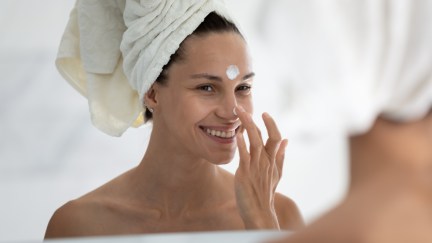 A woman puts sunscreen on her face while smiling in the bathroom mirror.