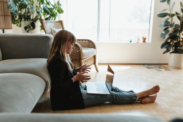 A woman leans against her couch on a web call with a laptop on her lap.