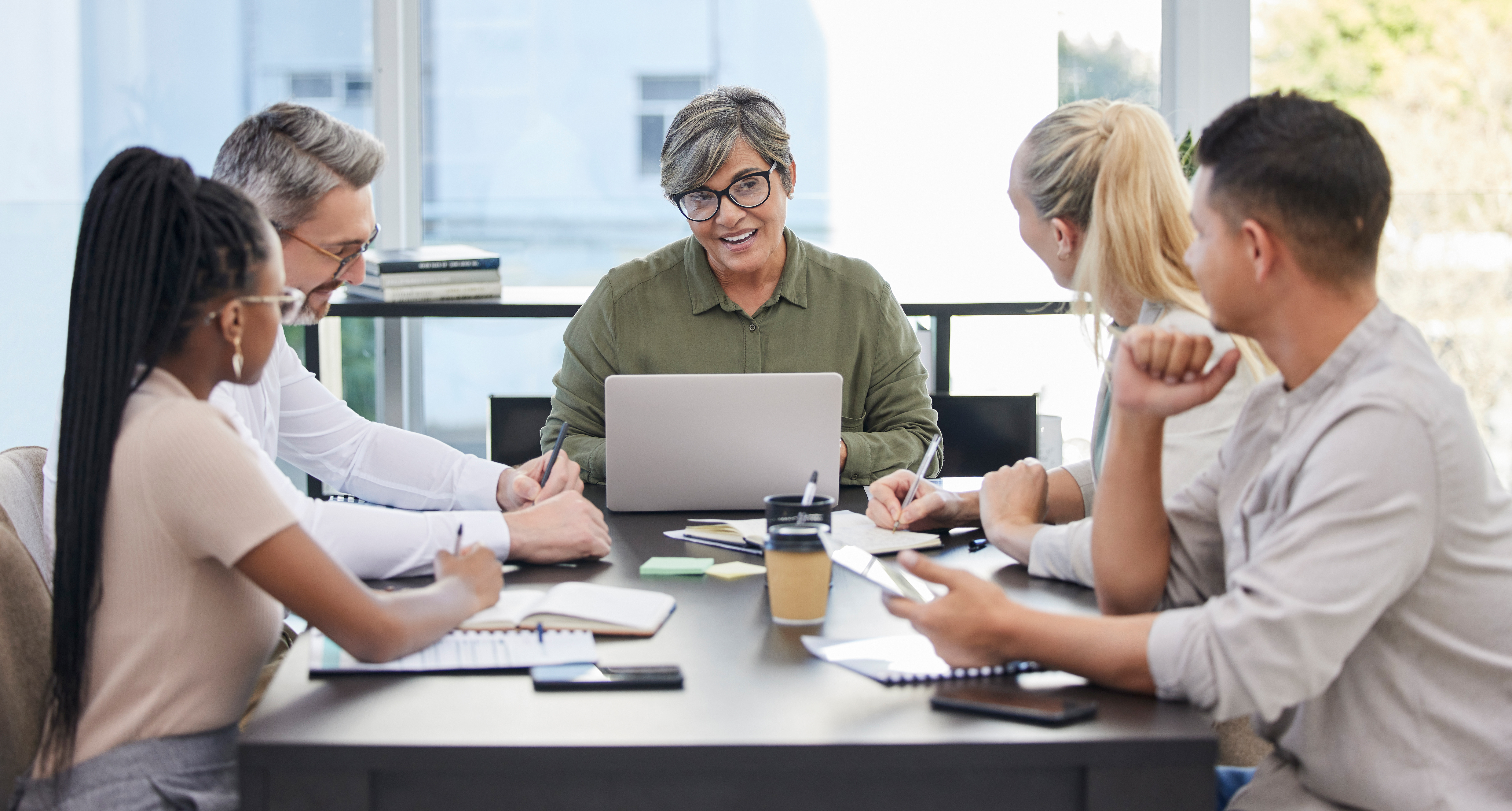 Older leader at head of table with laptop talking with group of younger employees