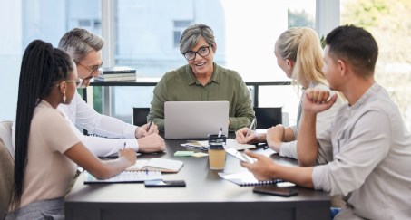 Older leader at head of table with laptop talking with group of younger employees