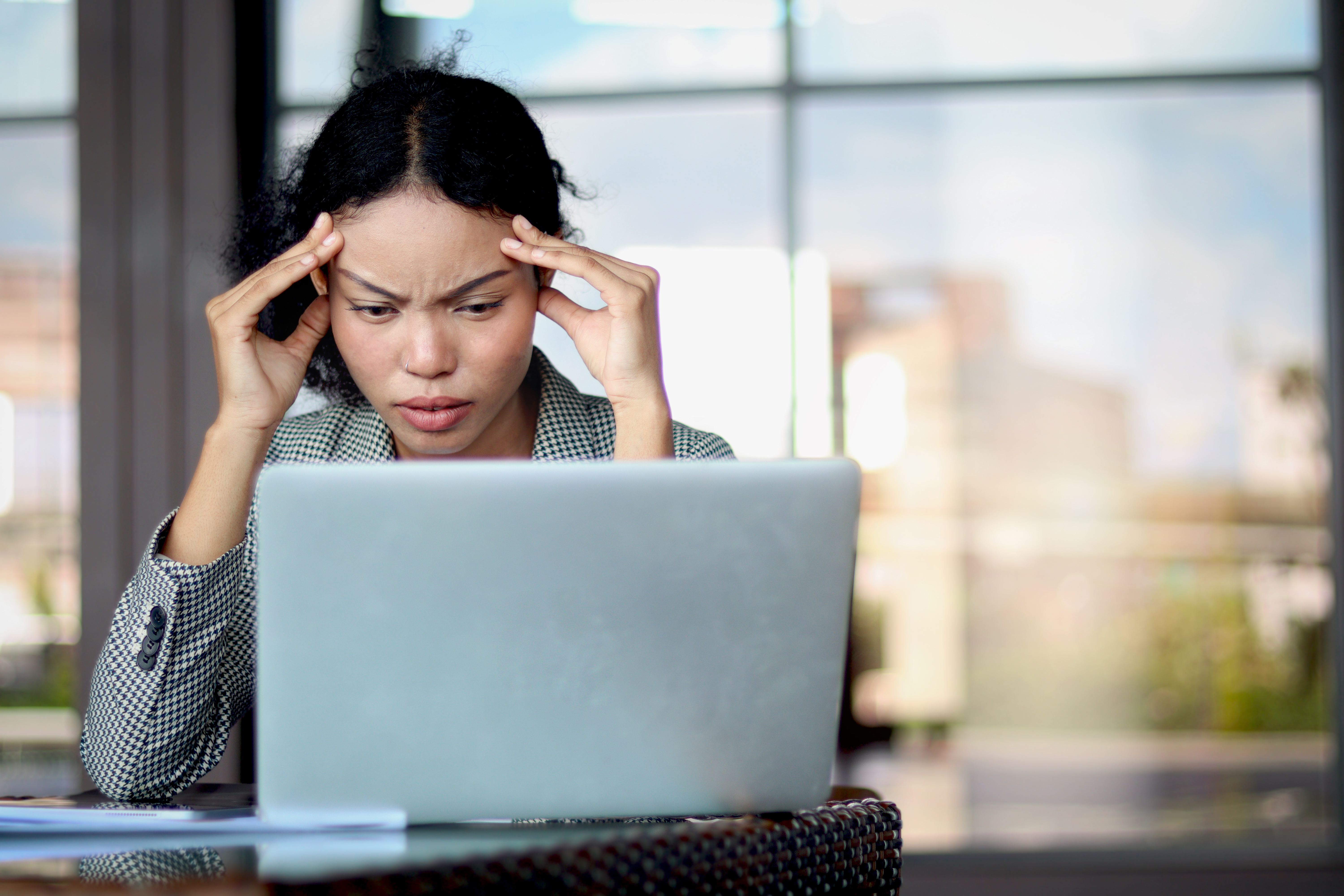 A woman rubs her temples while looking down at her laptop; she sits in front of large glass window.