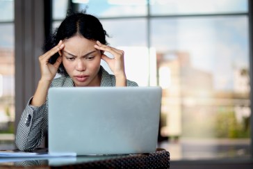 A woman rubs her temples while looking down at her laptop; she sits in front of large glass window.