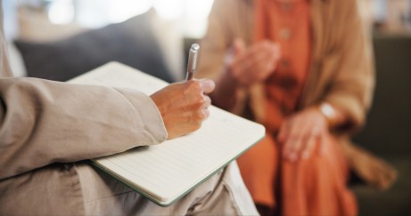 A woman writes on a notebook while another woman sits on the couch in front of her.