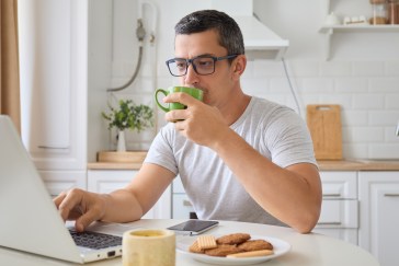 A man sips his coffee while typing on his laptop with one hand; his breakfast sits untouched on the table.