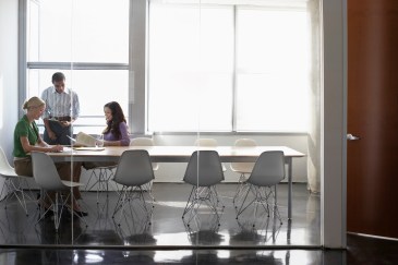 Three people sit in a conference room that's separated from the rest of the office by a floor-to-ceiling glass window.