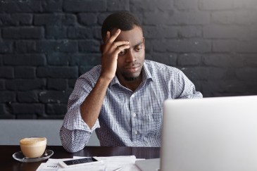 A man looks down at his laptop in frustration; a latte sits on the desk.