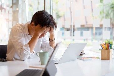 A young man has his head in his hands while looking down at his laptop.