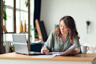 Woman writing on paper, open laptop on table in home