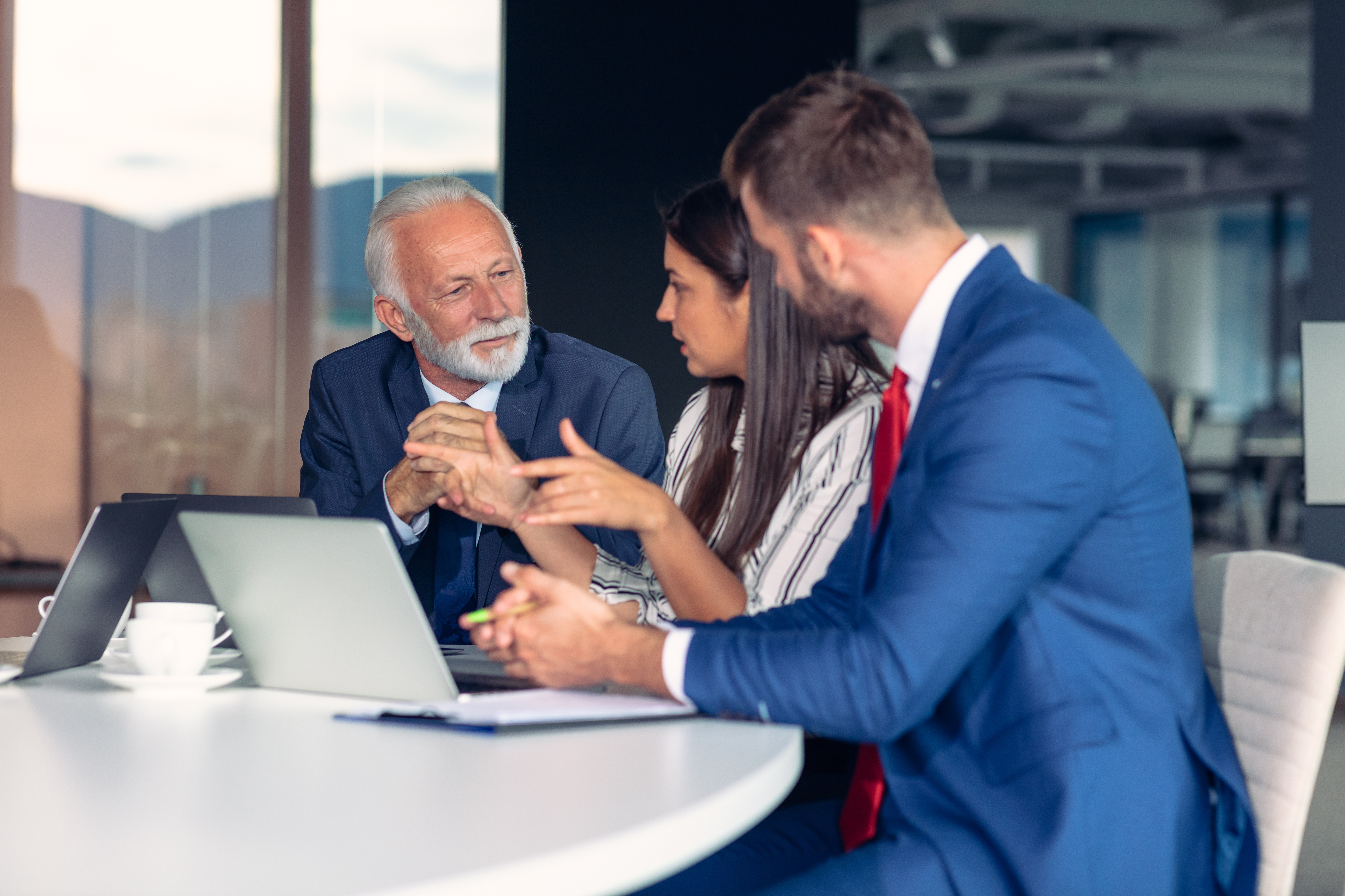 Older male employee and two younger employees sitting at table in office talking and working