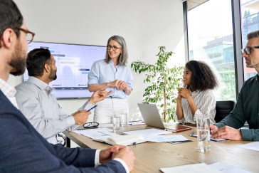Woman standing, leading meeting of people around table