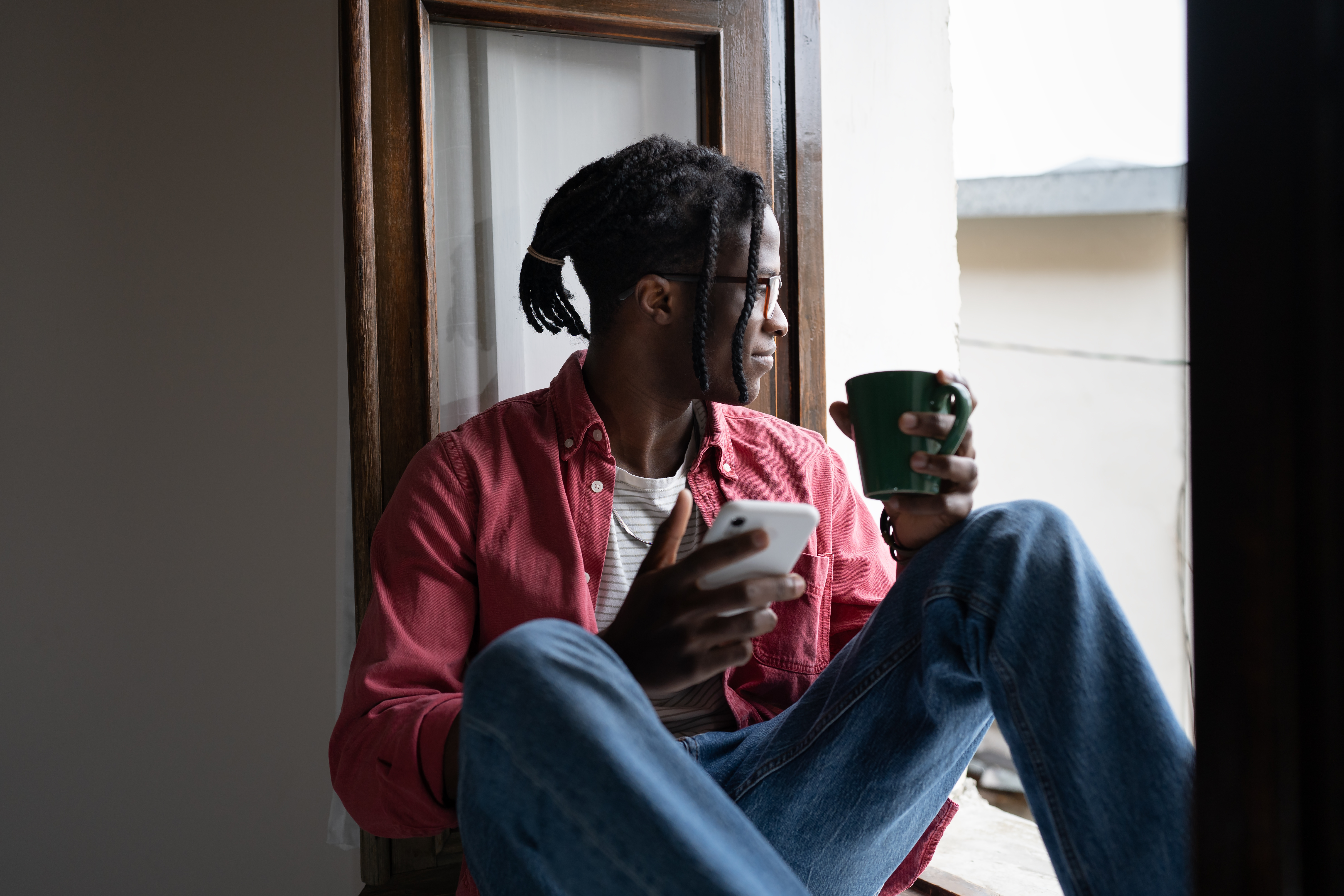 Man sitting on window sill looking outside with phone and coffee mug in hands