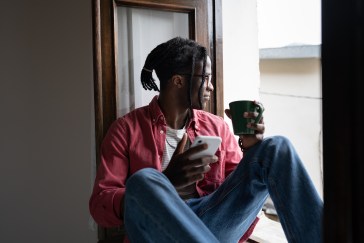 Man sitting on window sill looking outside with phone and coffee mug in hands