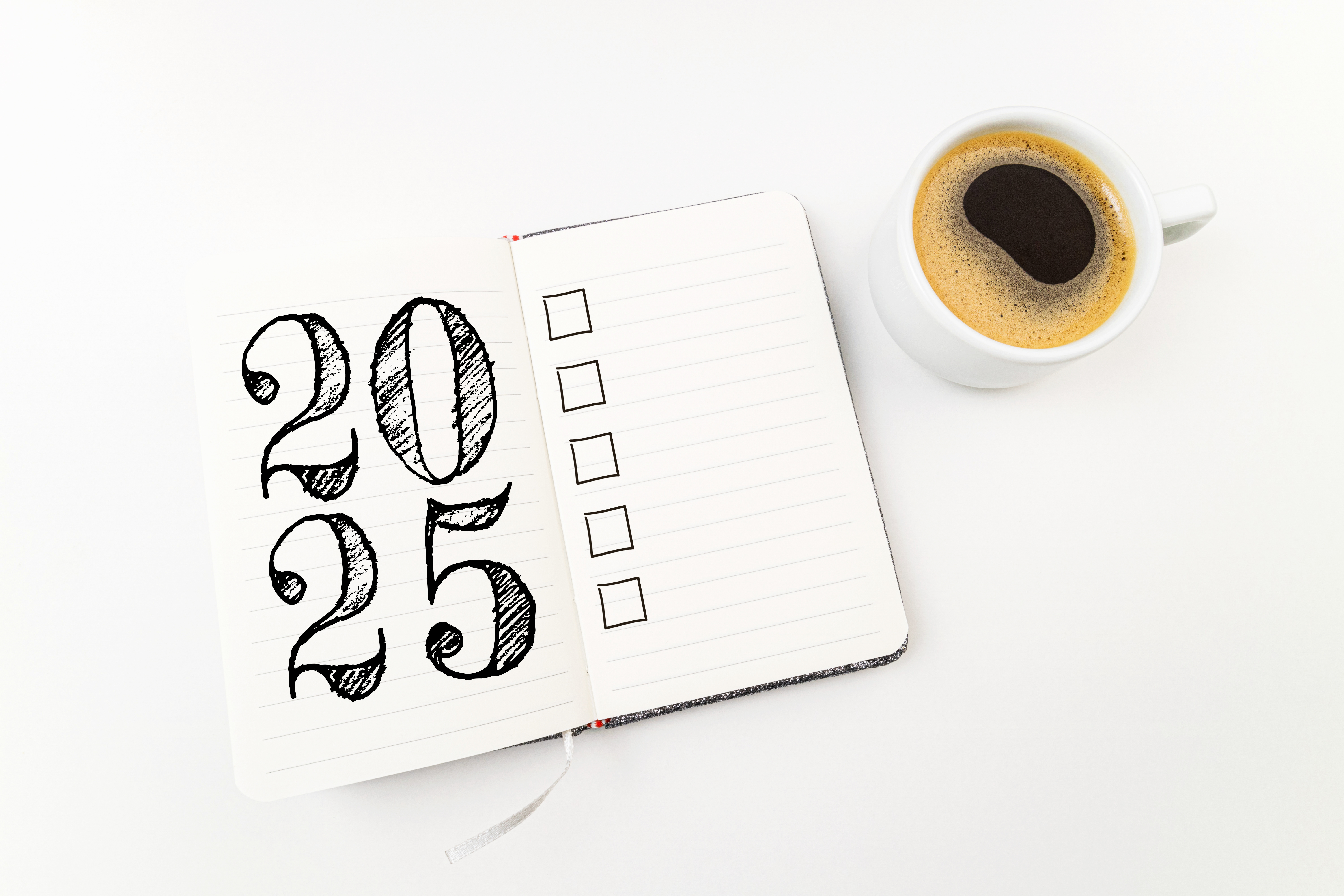 A notebook is open on an all-white table and says "2025" in big black letters. Next to it, there's a cup of coffee.