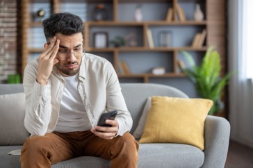 A man sits on his couch, looking disappointed at his phone.