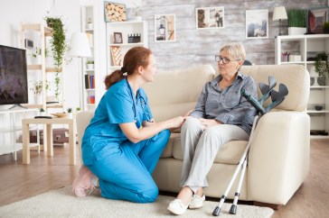 Medical professional helping woman sitting on couch with crutches