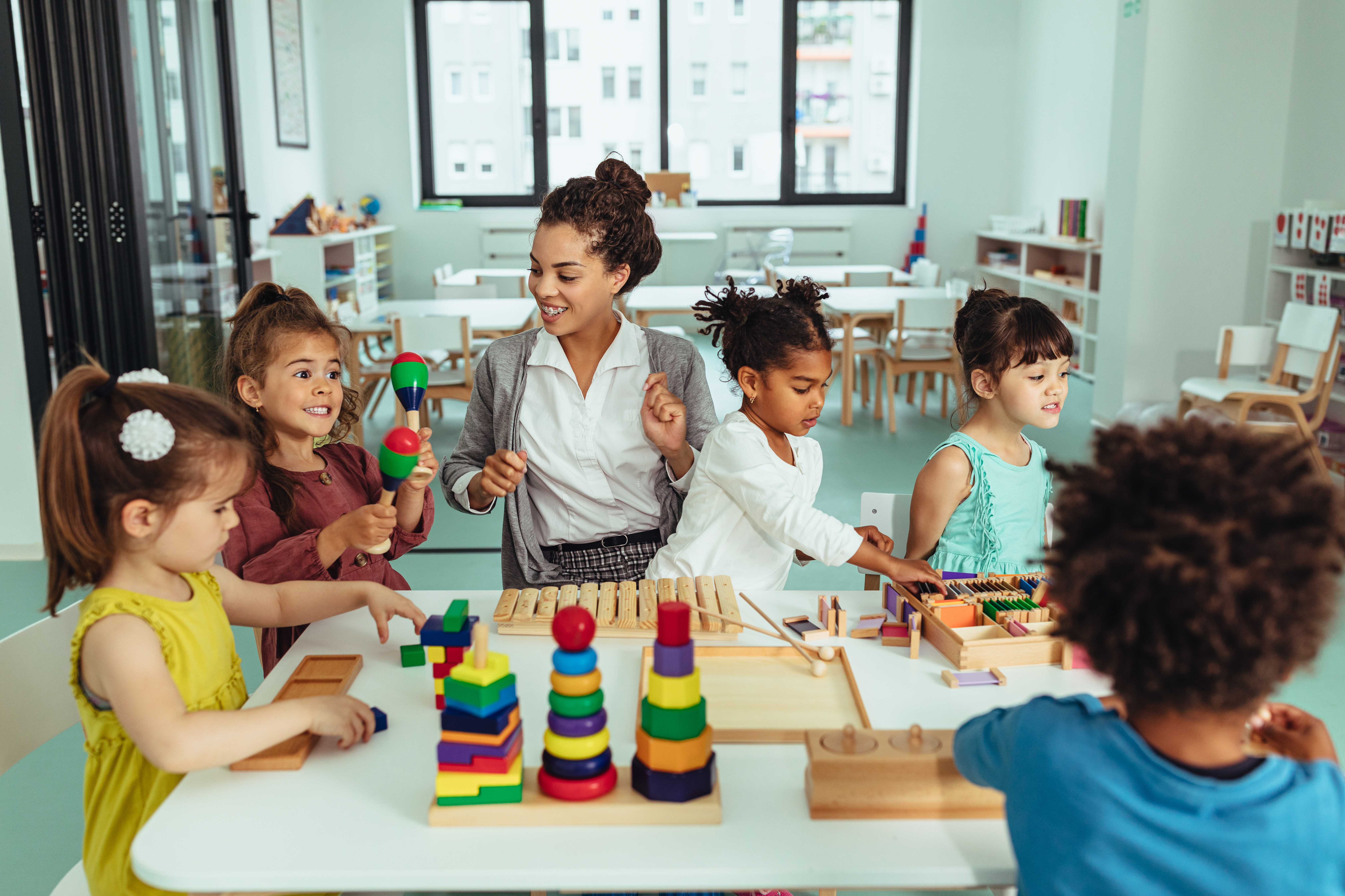 A group of children play with toys at a table alongside their preschool teacher.