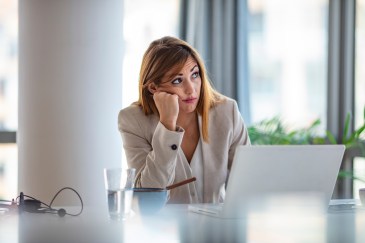 Woman in suit sitting at desk in office with head on first, looking frustrated, confused