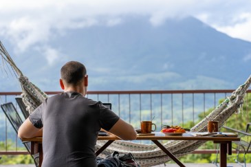 A man sits on a patio, working on his laptop, with a view of mountains and clouds drifting over them.