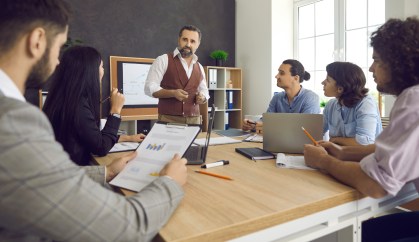 Group of employees sitting around table listening to man standing