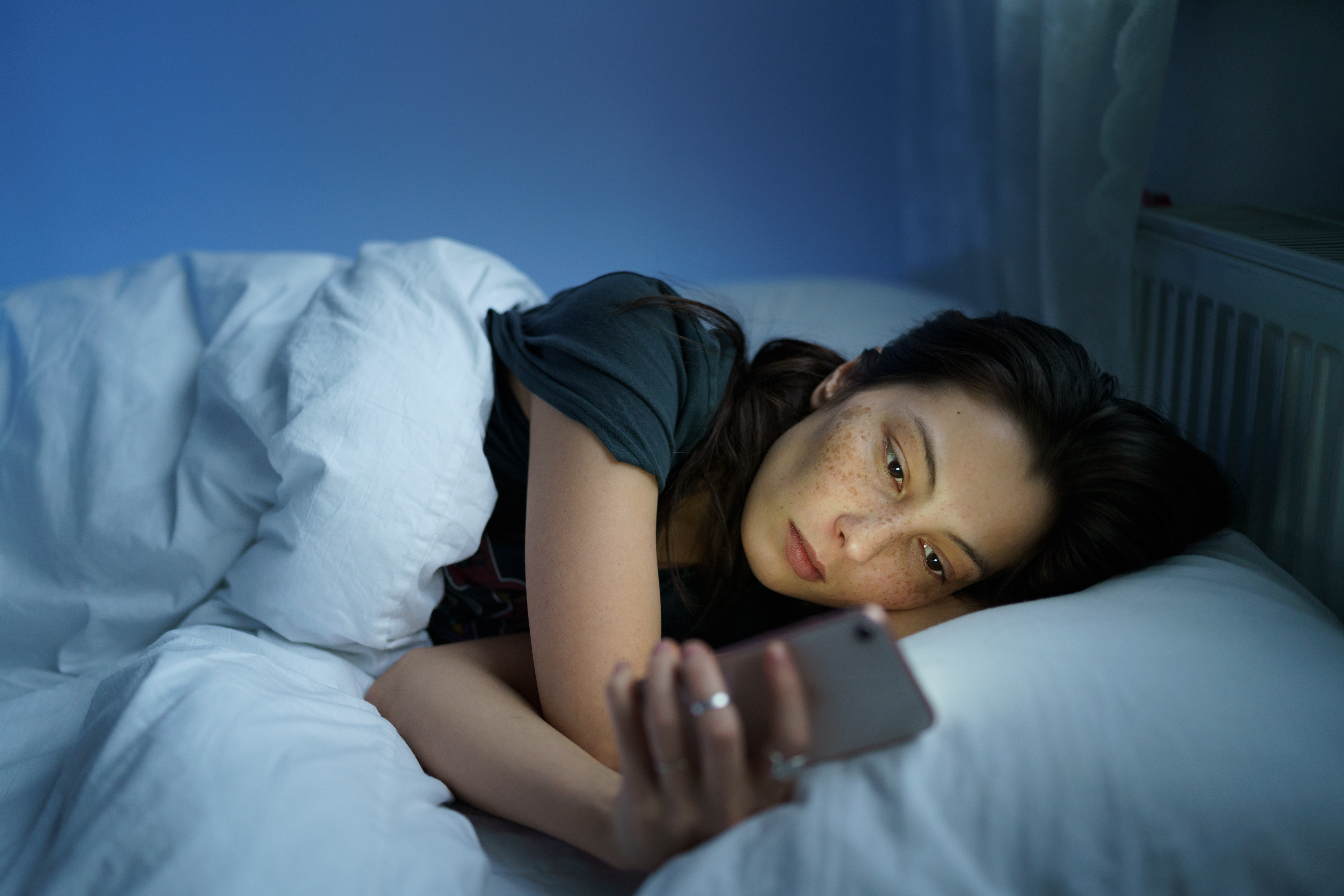 A woman stares at her phone while laying in her bed.