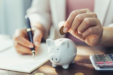 Woman's hand putting coin into piggy bank and writing into a notebook