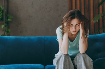 A young woman sits on a couch; she has her head in her hands in obvious discomfort.