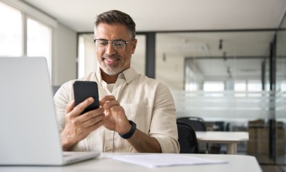 A middle-aged man is smiling at his phone while at his desk at work.