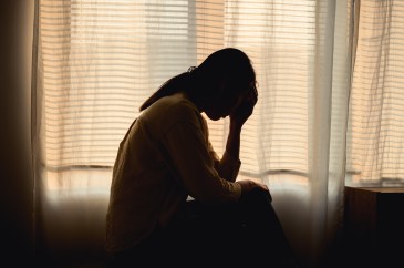 A woman silhouetted in a dark bedroom has her head in hands.