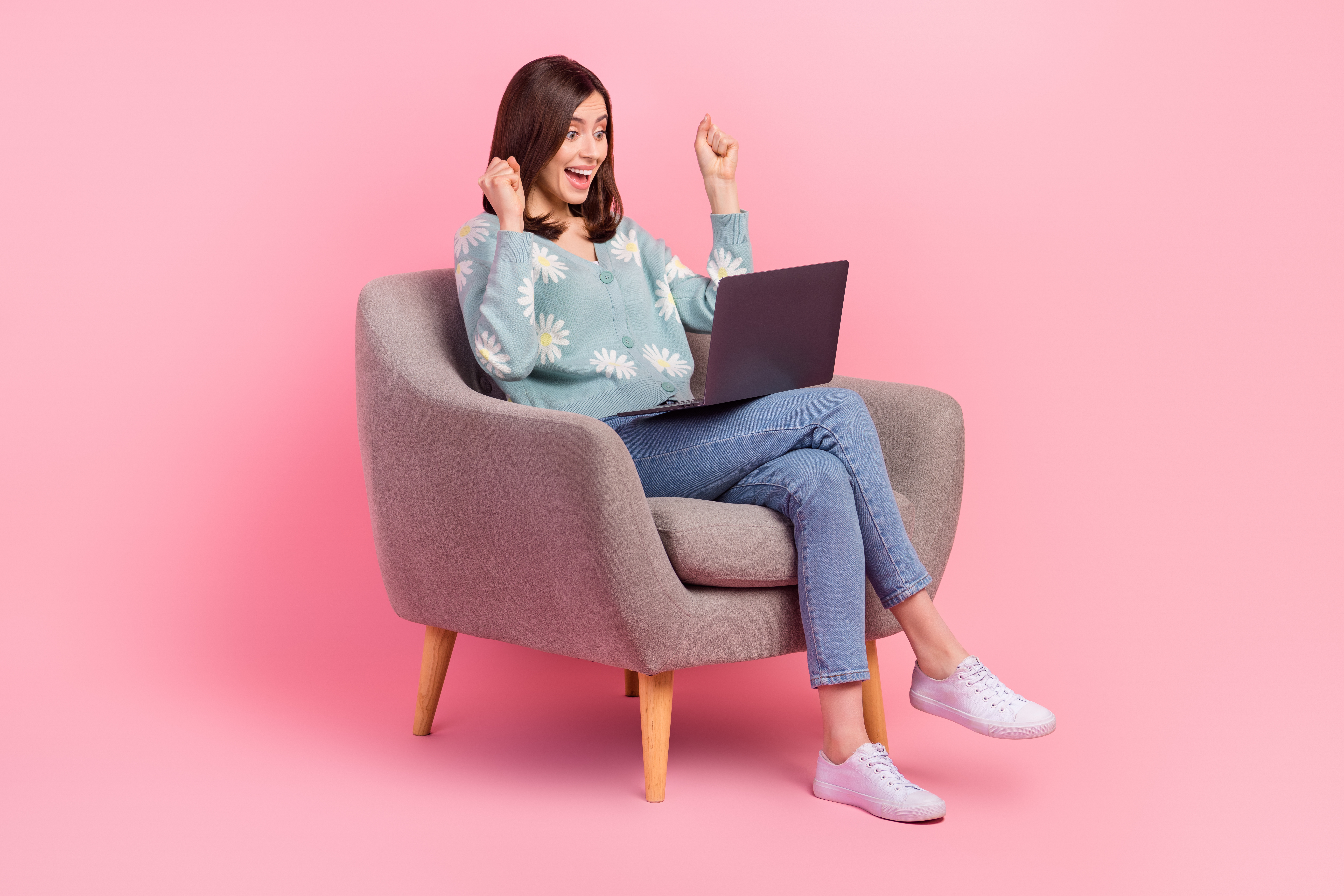 A woman sits on chair and pumps her fists in excitement while looking at her laptop.