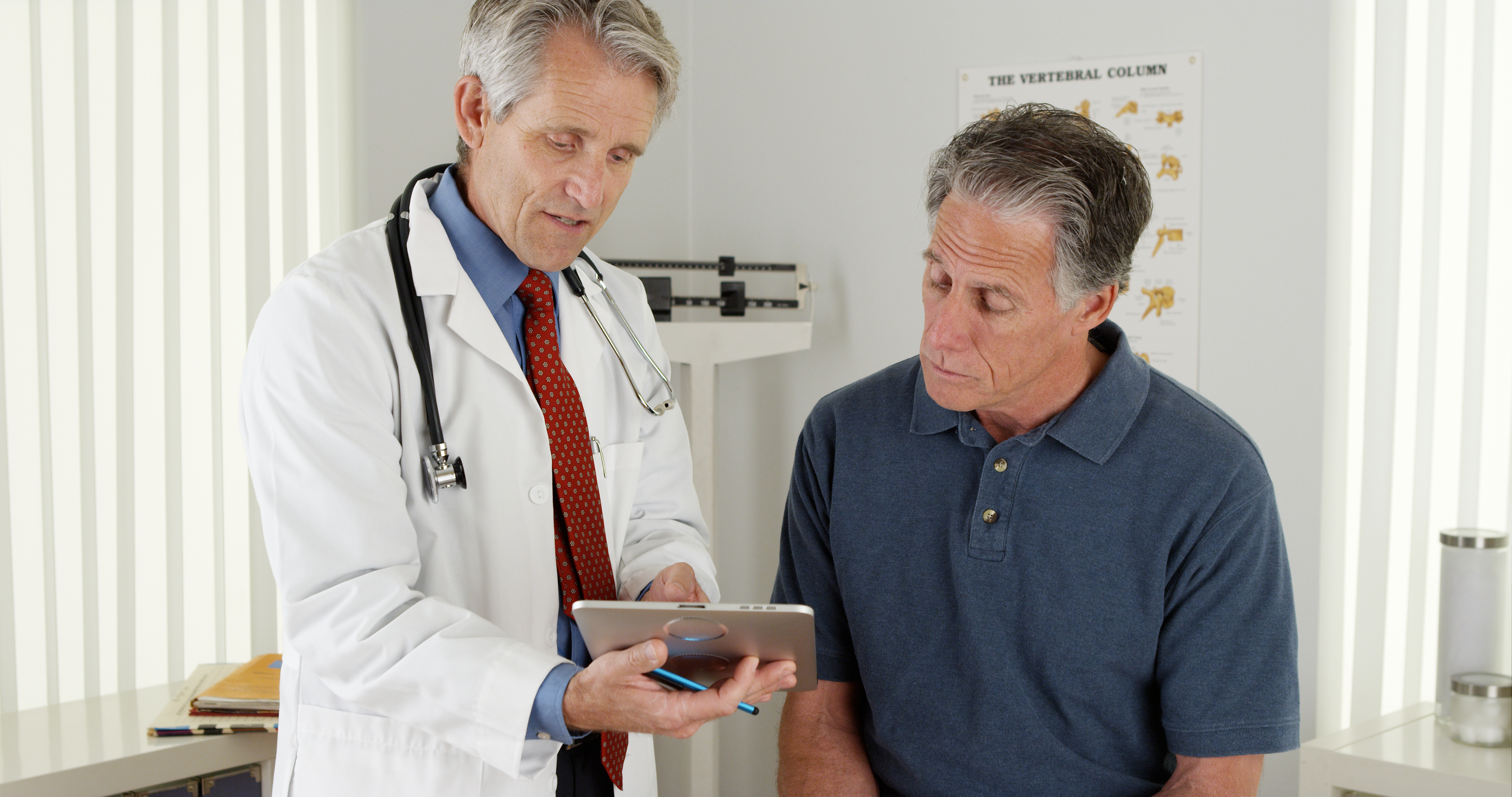 Male doctor talking to middle-aged male patient, showing him information on iPad.
