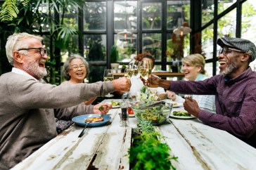 Old friends toast with champagne at a trendy restaurant with greenery in the background.