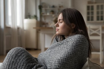 A young woman is wrapped in a blanket, sitting on the couch, her eyes closed in pain.