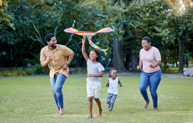 A Black family runs through a field with a colorful kite, laughing.