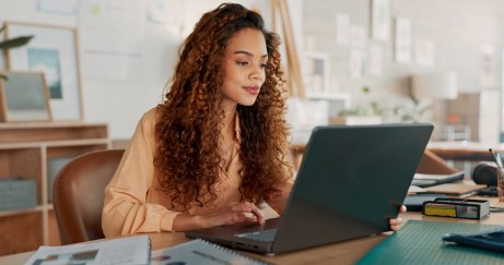 A woman looks at her laptop in her daylit home office.