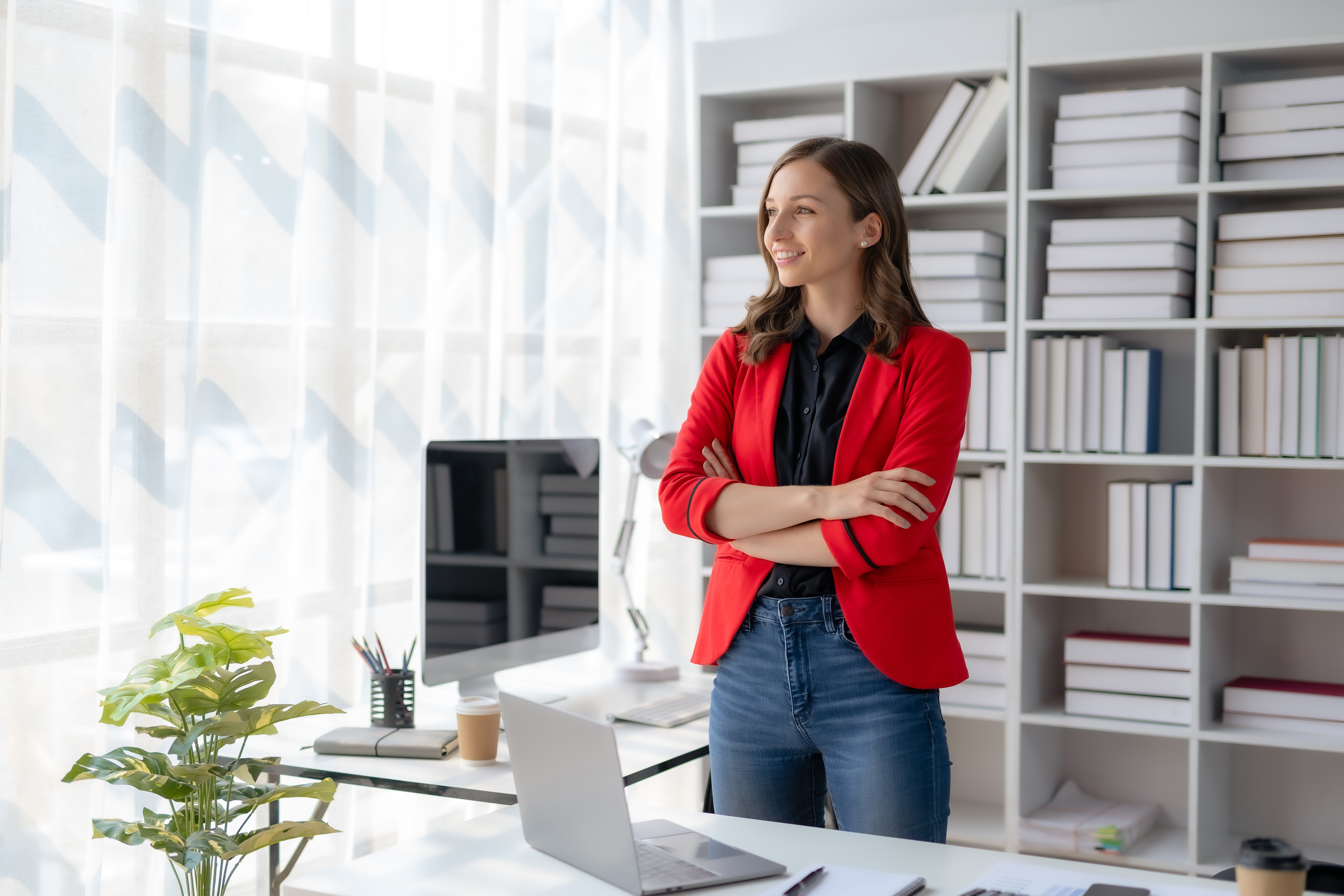 Young female employee in red blazer and jeans standing behind desk with laptop