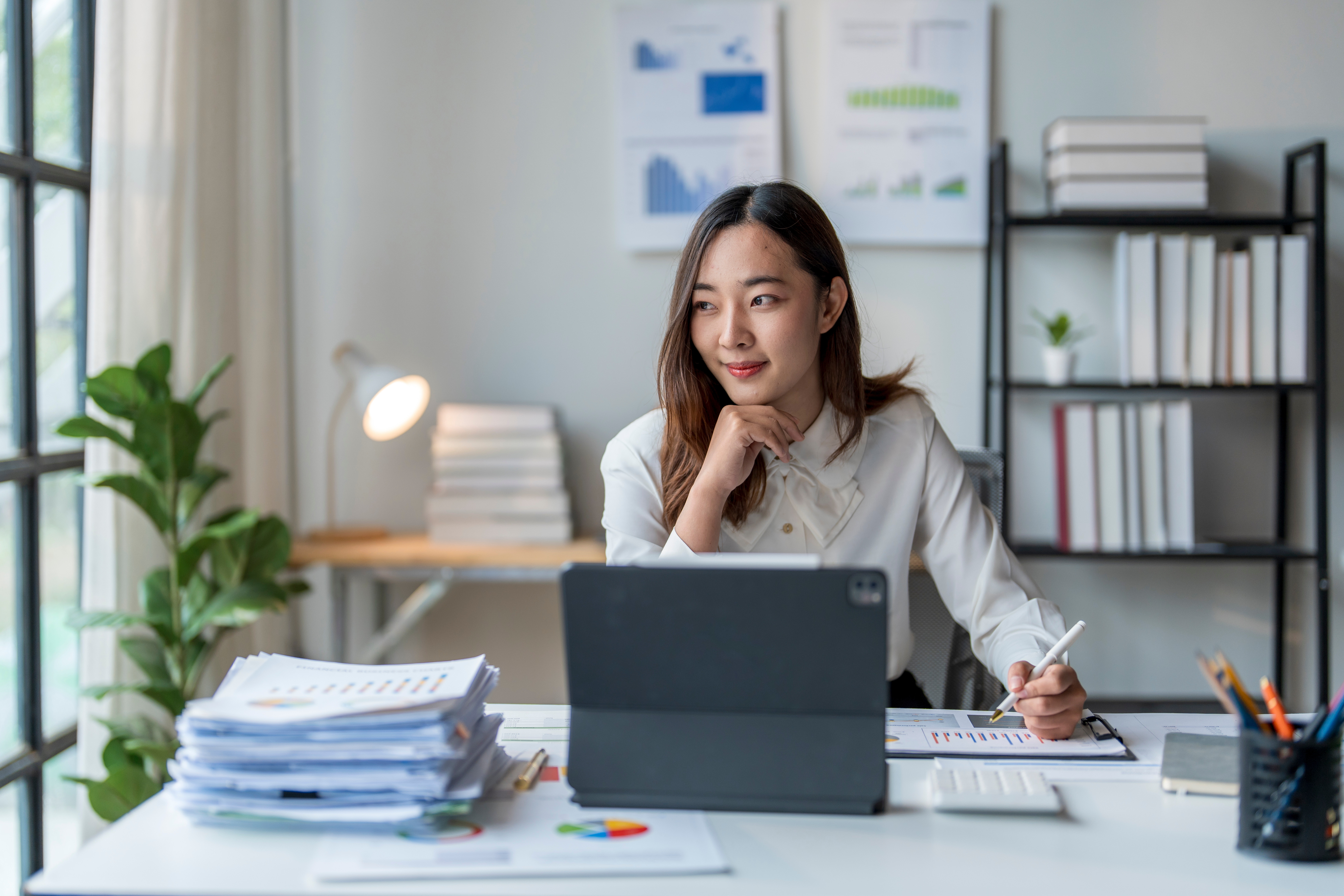 Woman sitting at desk with laptop and paperwork, looking out window, smiling