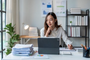 Woman sitting at desk with laptop and paperwork, looking out window, smiling