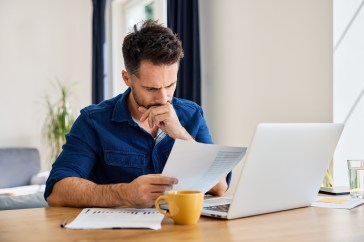 Man looking intently at paperwork, sitting at table with laptop