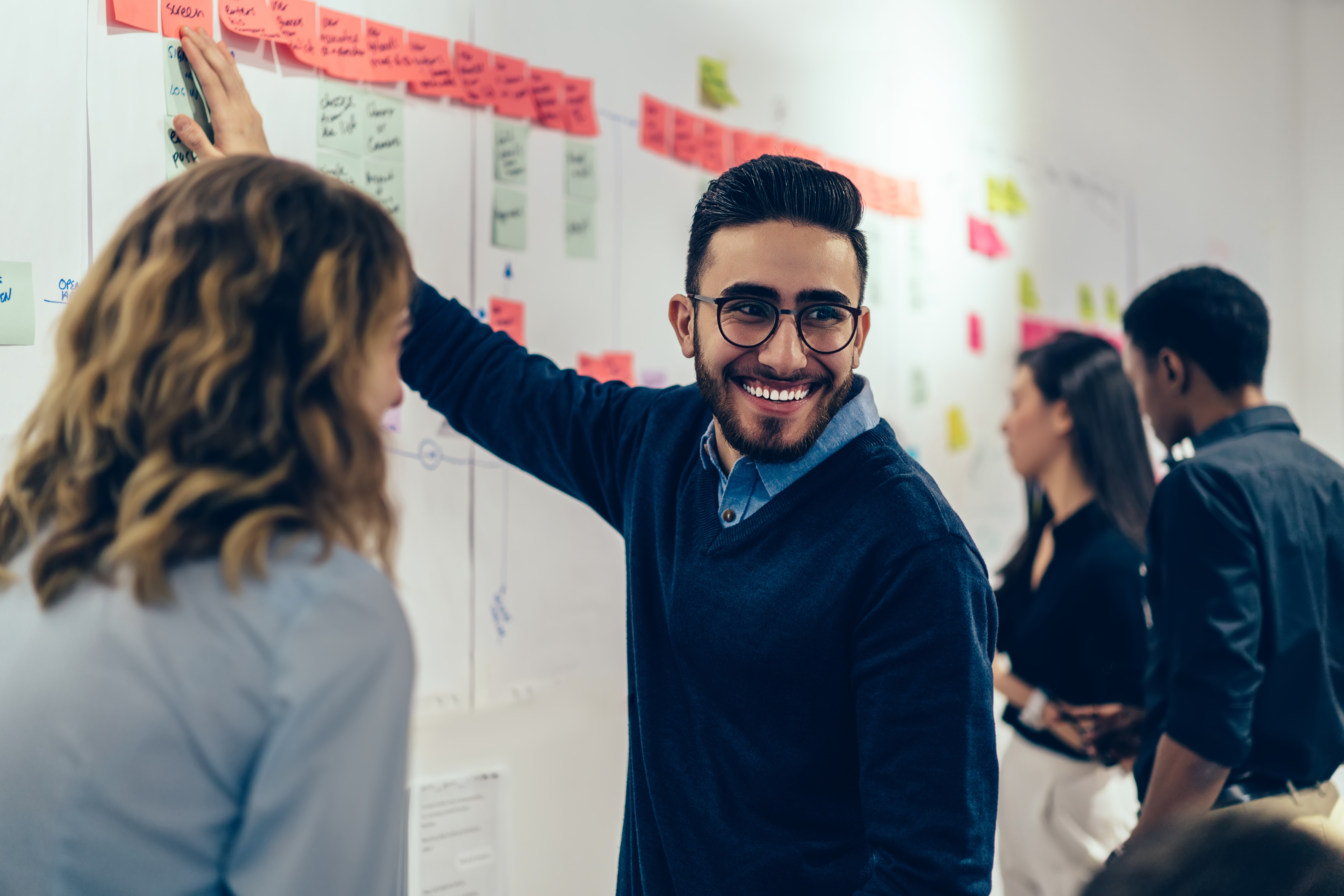 Male employee pointing to whiteboard with sticky notes, smiling, female employee next to him