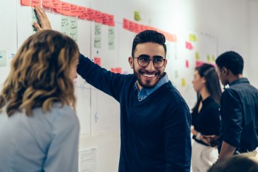 Male employee pointing to whiteboard with sticky notes, smiling, female employee next to him