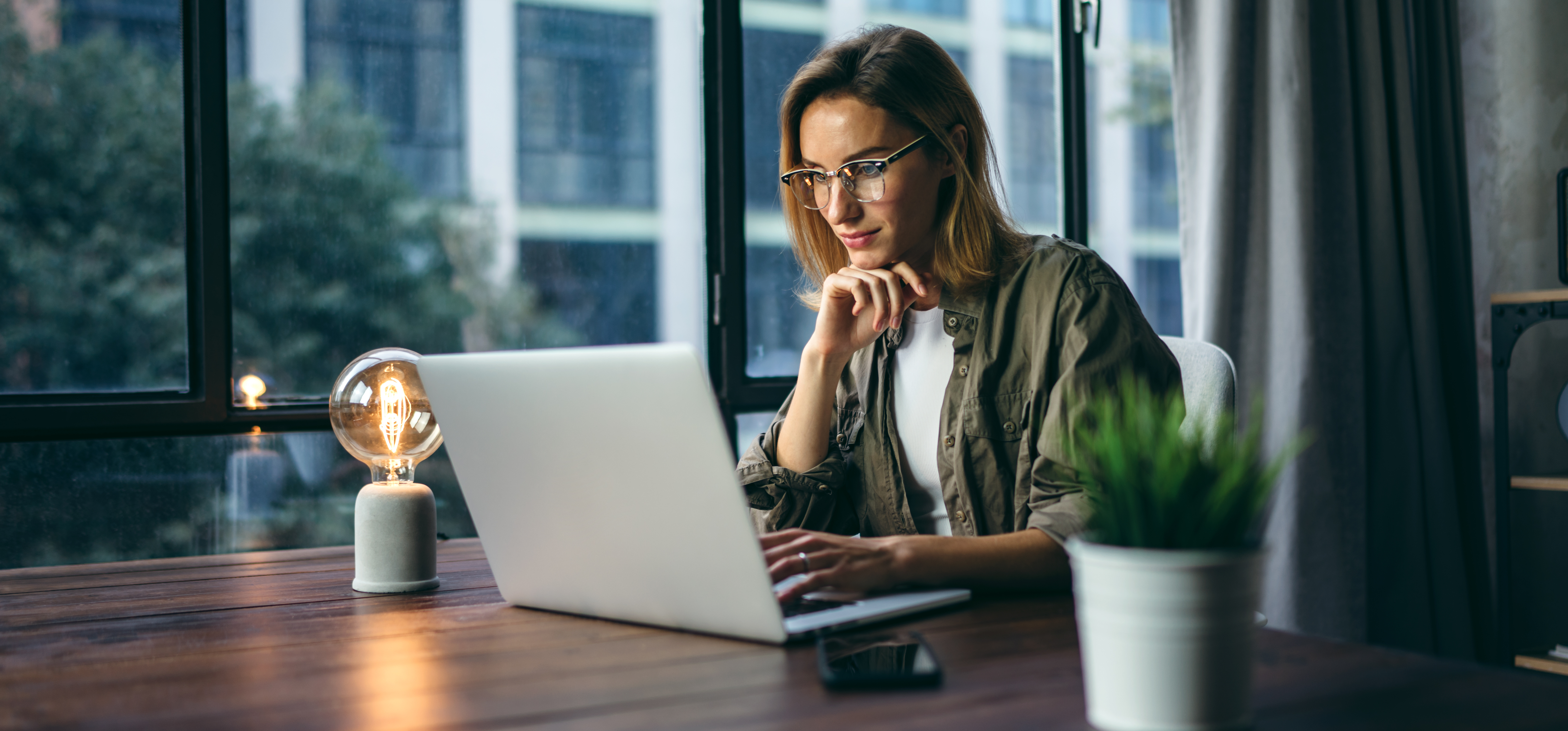 Woman working on computer, wearing glasses, sitting at desk with plant and lamp