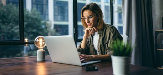 Woman working on computer, wearing glasses, sitting at desk with plant and lamp